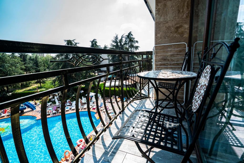 Balkons ar skatu uz baseinu, kur atpūšas cilvēki, un apkārt aug koki. Uz balkona ir melna metāla galda un krēslu komplekts.