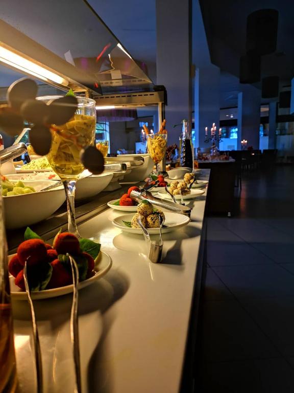 Bagātīgs bufetes galda klājums ar dažādiem salātiem un desertiem, tostarp augļu un saldējuma porcijām, kas pasniegtas elegantos traukos. Apgaismojums rada patīkamu atmosfēru.