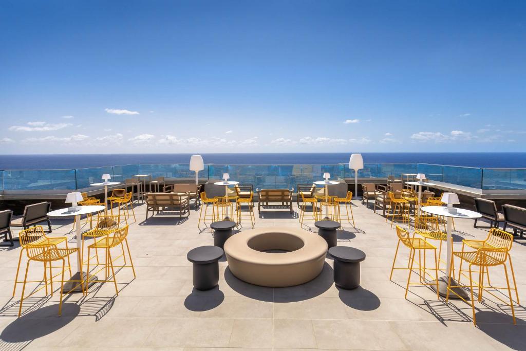 Mūsdienīgs terases bārs ar dzeltenām krēslām un baltām galdiem, kas piedāvā skatu uz jūru zem saules gaismas. Šī vieta ir ideāla relaksācijai un socializēšanai.