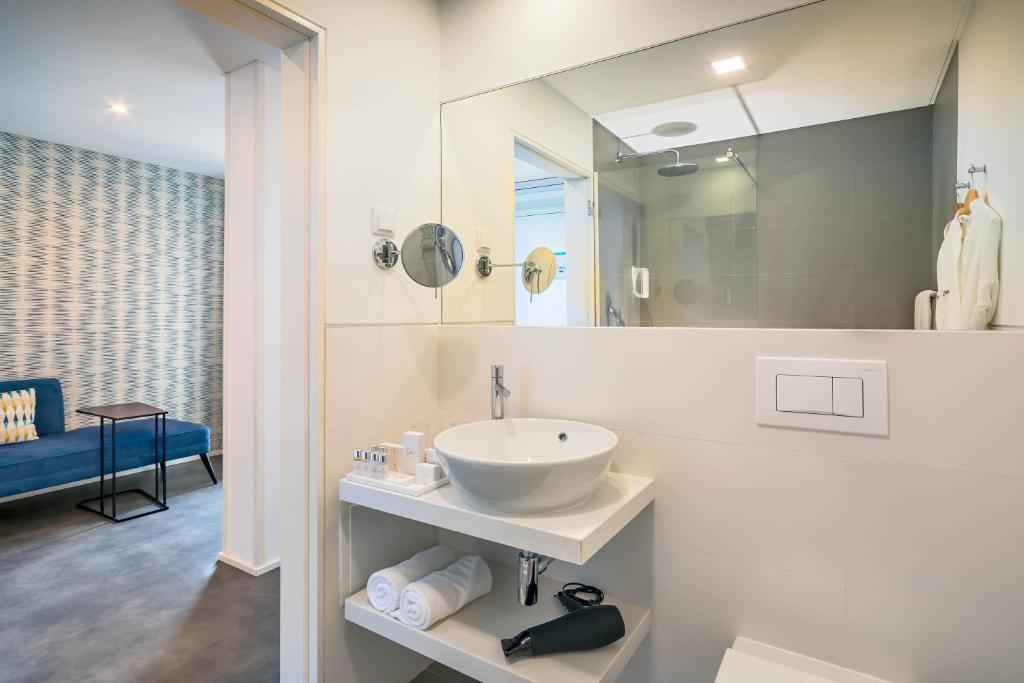 Mūsdienīga vannas istaba ar elegantu izskatu, kas ietver balto izlietni uz plaukta, dvieļus un matu žāvētāju. Fons ar dīvānu un stilīgu tapeti papildina telpas estētiku.