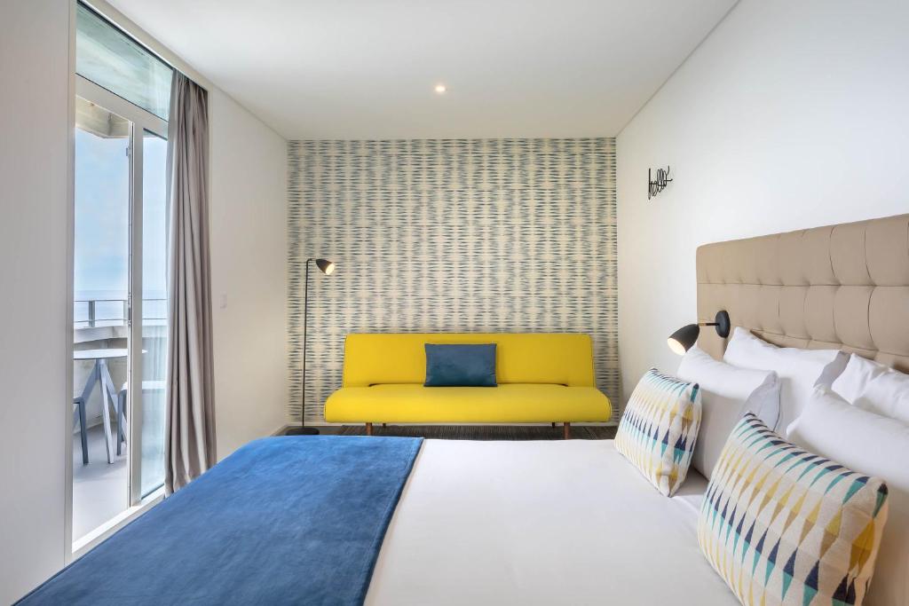 Mūsdienīga viesnīcas istaba ar gaišām krāsām, dzeltenu dīvānu un zilu gultas pārklāju, radot relaksējošu atmosfēru. Logu caurspīdīgums nodrošina dabīgu apgaismojumu un piekļuvi balkona skatiem.