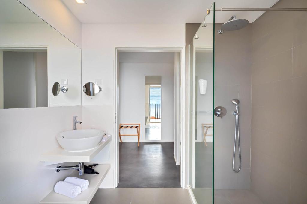 Mūsdienīga vannas istaba ar stikla dušu un minimālistisku dizainu, kurā ir baltas flīzes, izlietne un spogulis. Telpa ir gaiša un gaumīga, ar skatu uz ārpusi.