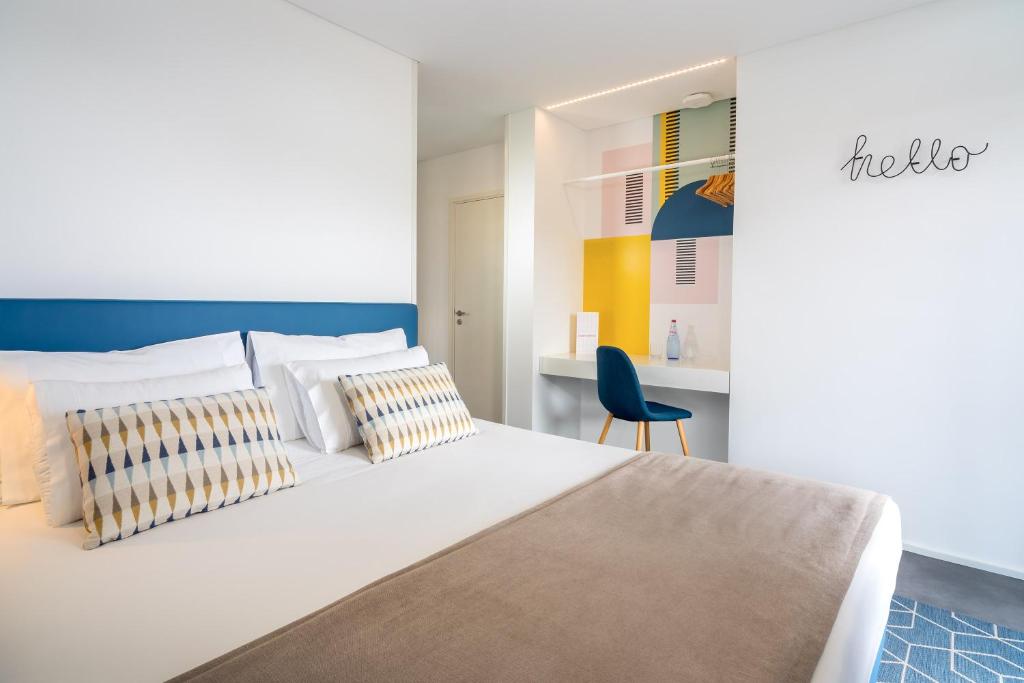 Mūsdienīga viesnīcas numura iekārtojums ar zilu gultas galvgali, dekoratīvām spilveniem un stilīgu darba zonu. Sienā ir krāsaina māksla un uzraksts 