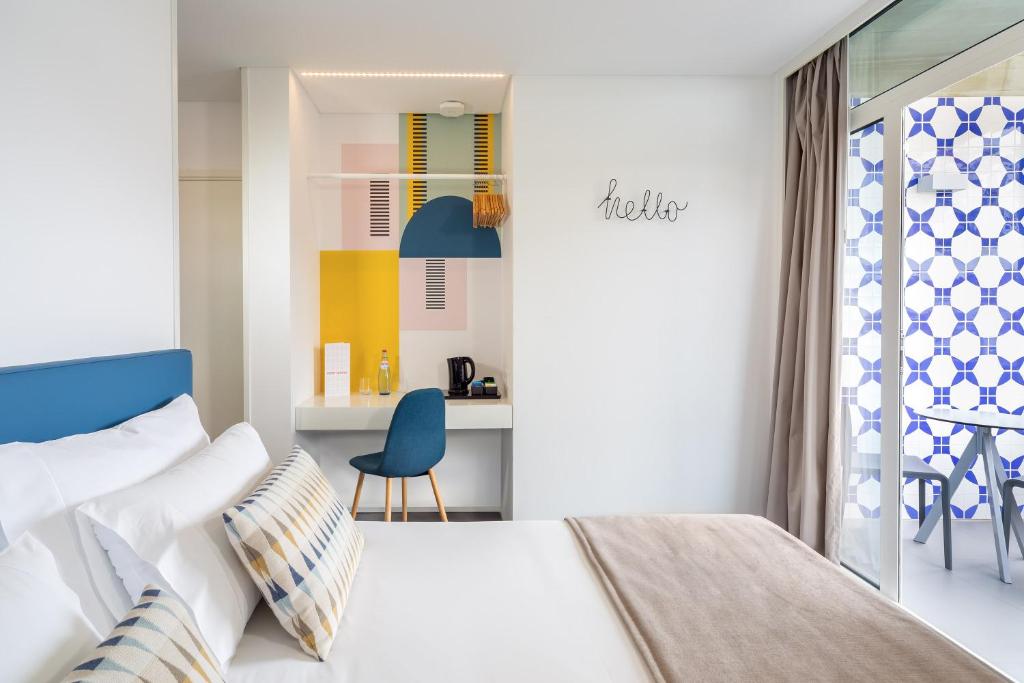 Moderns viesnīcas numurs ar gaišām krāsām un stilīgu dizainu, kurā redzams ērtā gulta, rakstāmgalds un logi ar dekoratīvām žalūzijām.