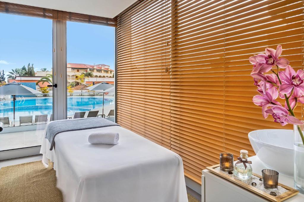 Relaksējoša spa telpa ar skatu uz baseinu, kur atrodas masāžas galds un dekoratīvas orhidejas. Ideāla vide atpūtai un labsajūtai.