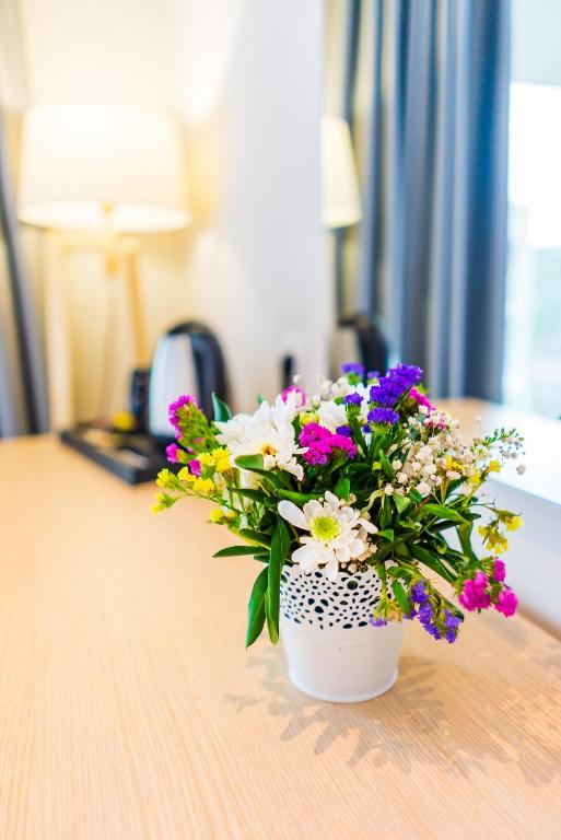 Krāsains ziedu pušķis baltā podiņā, novietots uz koka galda, blakus lampai un tējkannai. Ideāla interjera dekorācija, kas piešķir mājīgumu telpai.