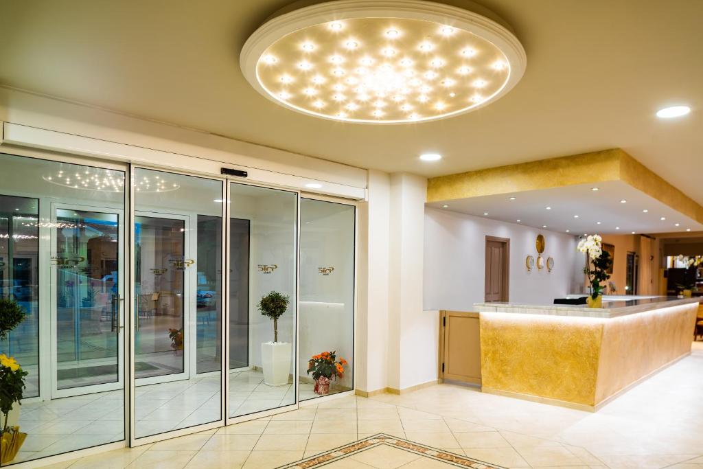 Mūsdienīgs viesnīcas vestibils ar stikla durvīm un elegantu apgaismojumu. Interjerā redzami dekoratīvi augi un stilīgs reģistratūras galds.
