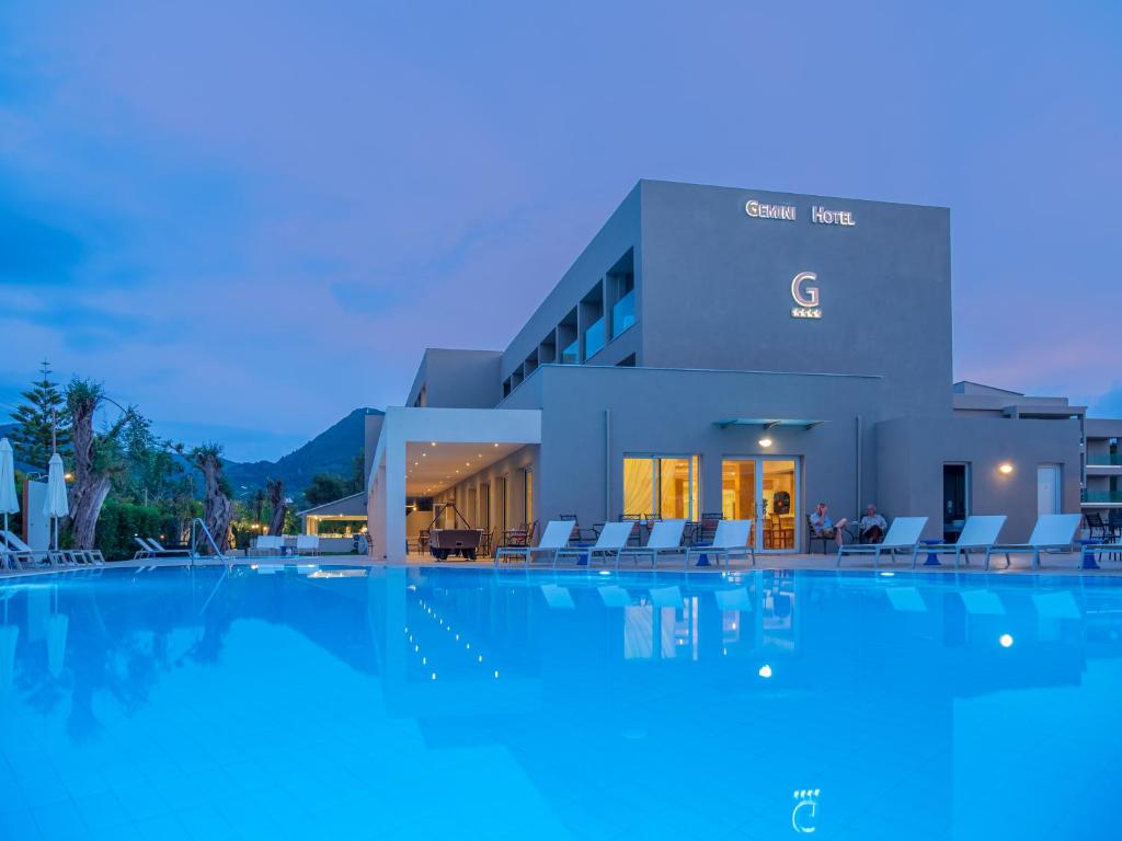Moderns viesnīcas ēka ar peldbaseinu un atpūtas zonu, iedegusies vakara gaismā. Viesnīcas nosaukums ir redzams uz fasādes.