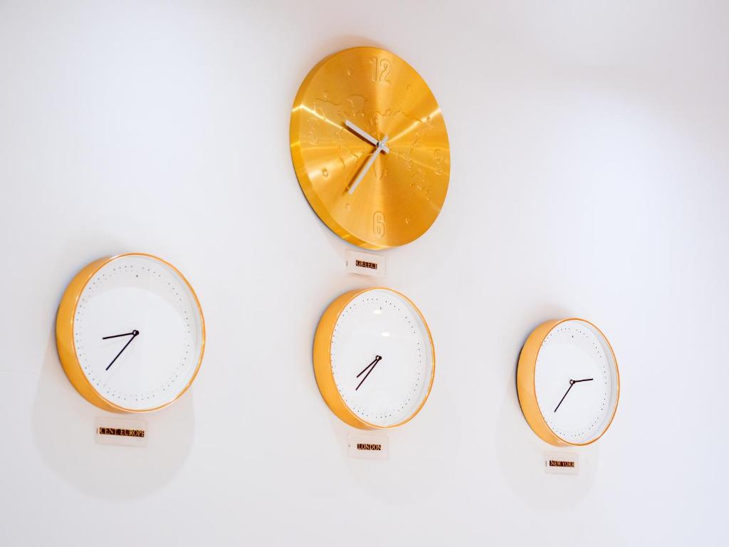 Trīs moderni pulksteņi ar dzeltenām apmalēm pie sienas, kas rāda dažādas laika zonas. Augšpusē ir liels pulkstenis, bet apakšā divi mazāki.