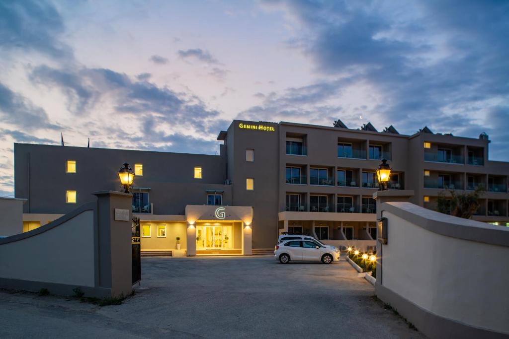Gemini viesnīca ar modernu arhitektūru un apgaismojumu, kas izceļas vakara laikā. Redzami balkoni un automašīna viesnīcas ieejā.