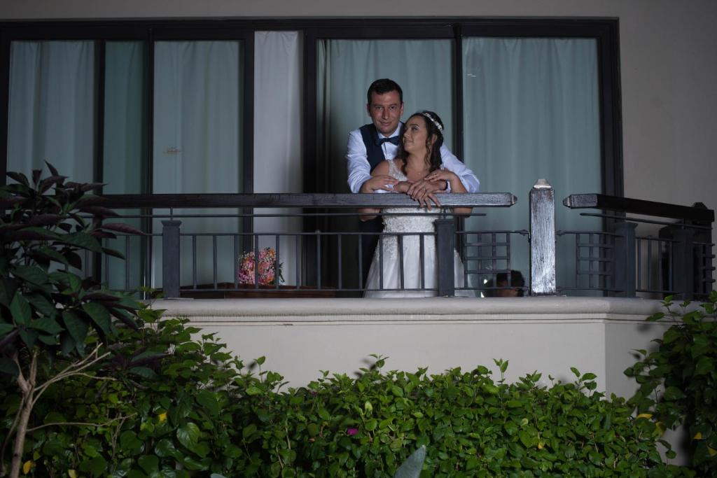 Jaunlaulātie stāv uz balkona, apskāvuši viens otru, tērpušies kāzās. Apkārt ir zaļa ainava ar augiem un ziediem, radot romantisku atmosfēru.