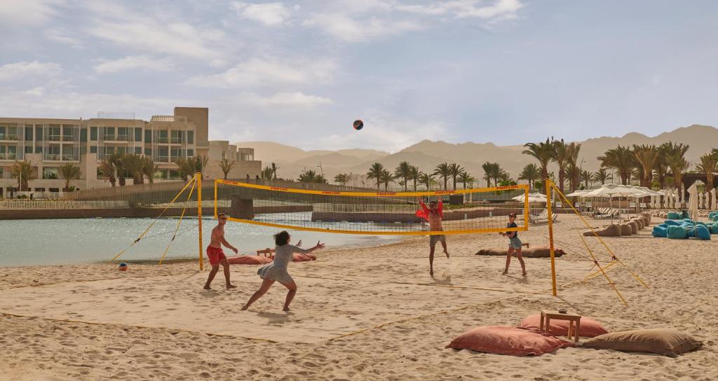 Pludmales volejbola spēles aina saulainā pludmalē ar palmām, fonā redzami viesnīcas un ērtas atpūtas zonas. Bērni izbauda aktīvu brīvā laika pavadīšanu.