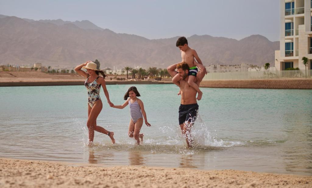 Ģimene izbauda laiku pludmalē, spēlējot ūdenī un smejoties. Fons veido kalni un moderni ēku kompleksi.