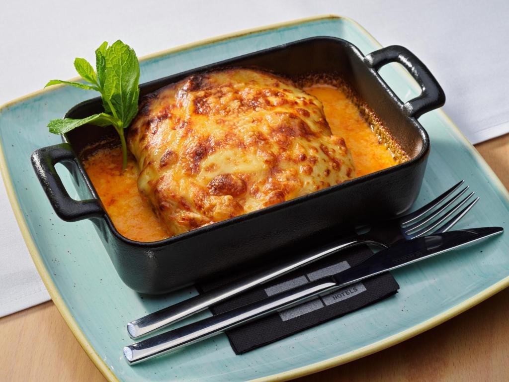 Sirsnīga lasagne panna ar zeltainu siera garoziņu, pasniegta melnā traukā uz gaiši zila šķīvja. Garnējums ar svaigu piparmētru lapu.