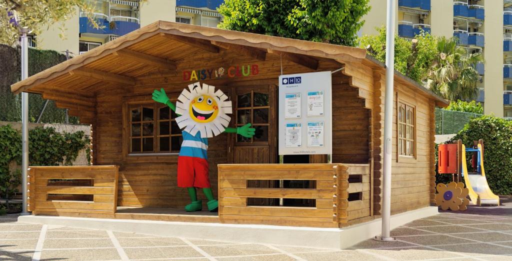 Koka māja ar uzrakstu "Daisy Club", kurā stāv jautrs un krāsains tēls ar smaidu. Apkārt ir bērnu rotaļu laukums un zaļumi.
