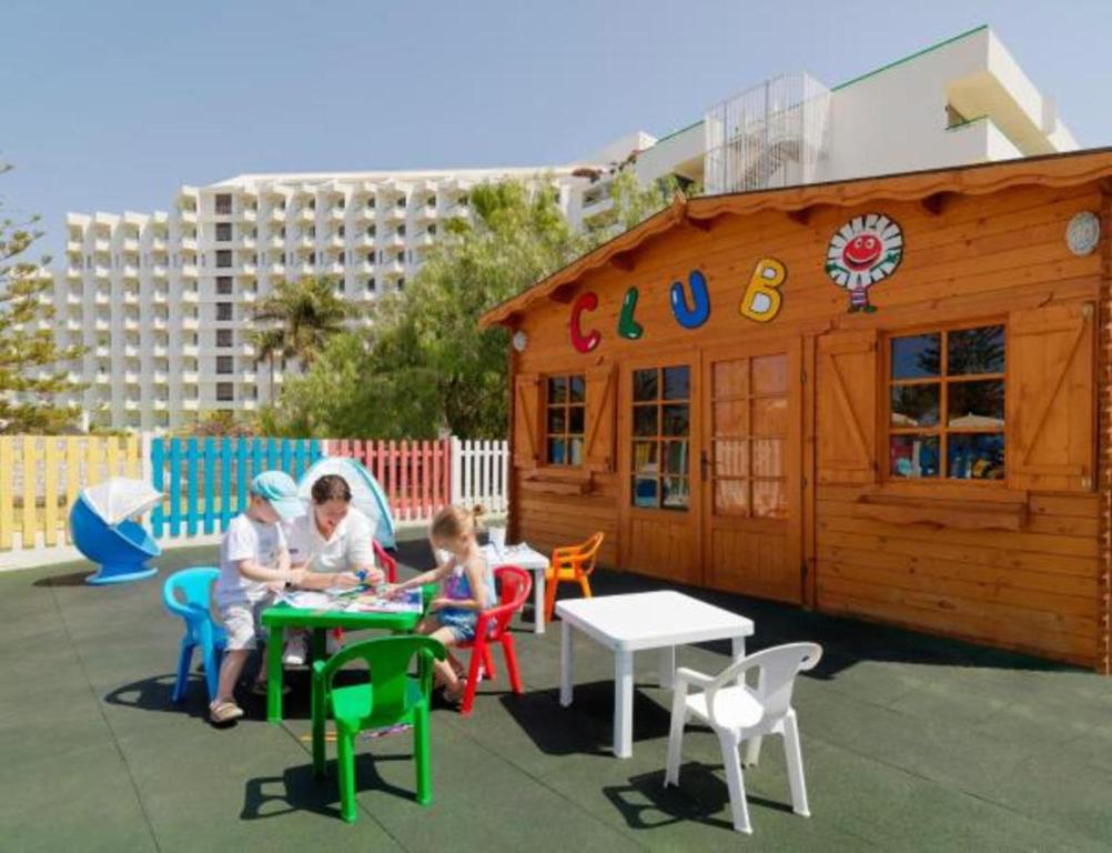 Bērnu klubs ar koka mājiņu un krāsainiem galdiem, kur bērni zīmē un spēlējas. Apkārt ir zaļa zāle un saulaina debess, radot priecīgu atmosfēru.