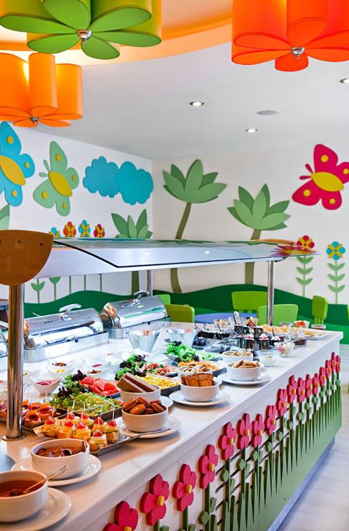 Krāsains bufetes galds ar dažādiem ēdieniem un dzērieniem, izvietots telpā ar dekoratīvām ziedu un tauriņu sienām. Gaismas piekari un spilgts interjers rada jautru atmosfēru.