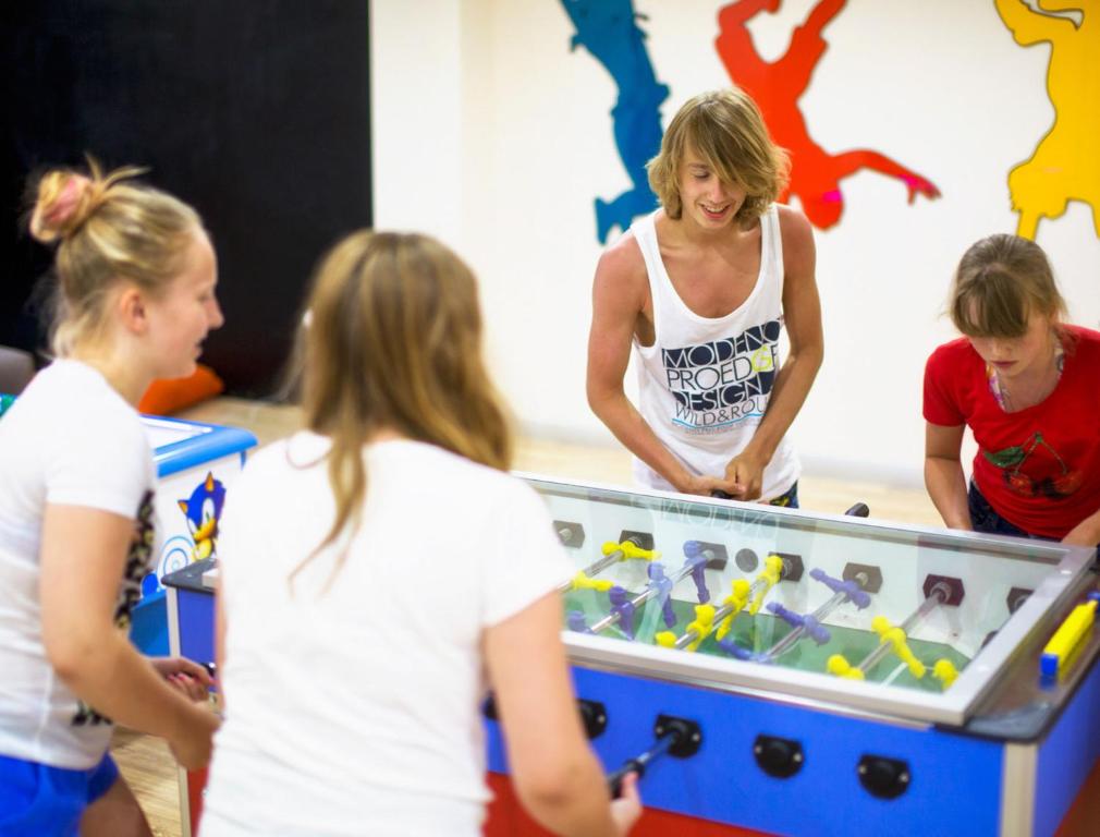 Jaunieši spēlē galda futbolu, izbaudot laiku kopā draugu lokā. Fons ir krāsains, ar dinamiskām figūrām uz sienas, kas uzsver izklaides atmosfēru.