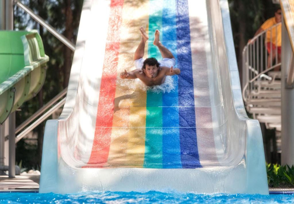 Bērns slidinās pa krāsainu ūdens slidkalniņu, baudot jautrību ūdens parkā. Fons ir zaļi koki un citi slidkalniņi, radot priecīgu atmosfēru.