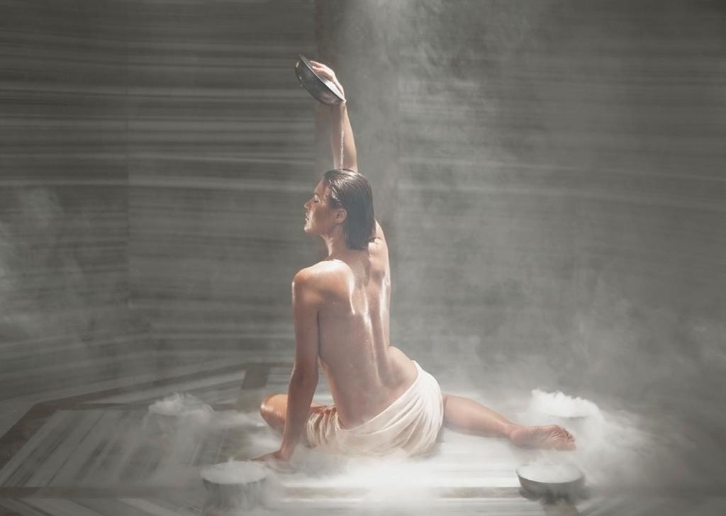 Sieviete sēž uz grīdas, izstiepjot roku ar trauku, tērpta vieglā drānā, apkārt ir tvaiks un gaisma, radot noslēpumainu atmosfēru.