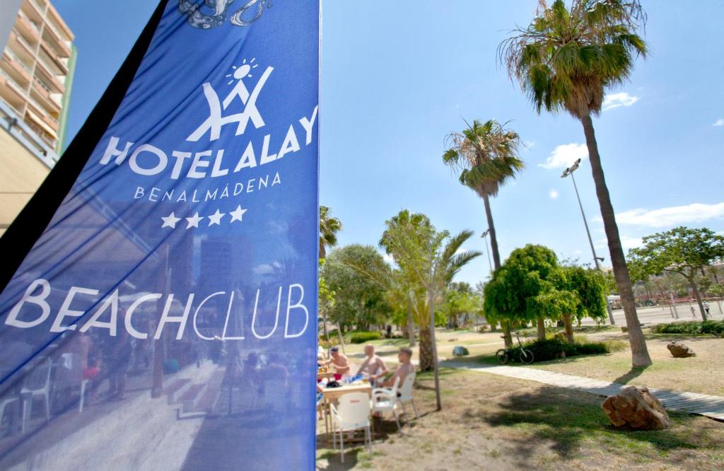 Zils karogs ar "Hotel Alay" un "Beach Club" uzrakstiem, fonā palmām un saulainu pludmales vidi Benalmādenā.