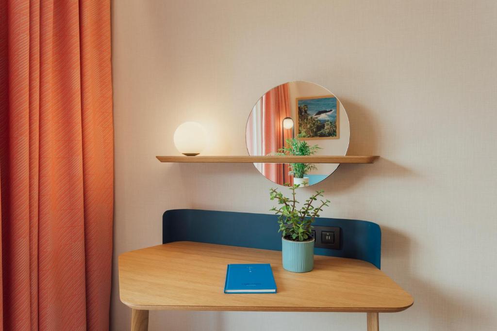 Mūsdienīga istabas stūra ar koka galdu, apgaismojuma lampu un spoguli. Uz galda atrodas zieds un zila piezīmju grāmatiņa.