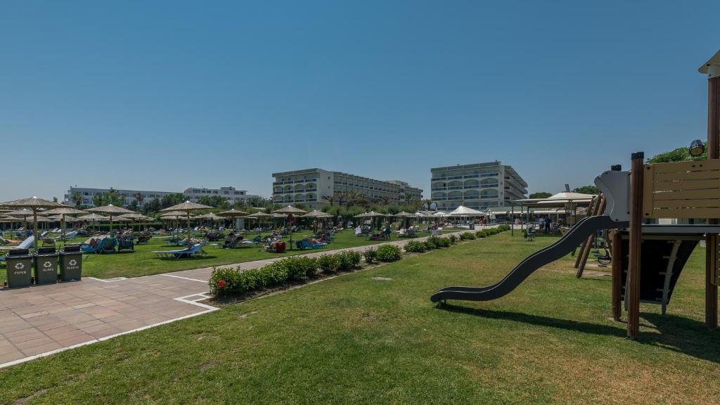 Plaša zāliena zona ar sauļošanās krēsliem, saulessargiem un bērnu rotaļu laukumu viesnīcas apkārtnē. Skats uz ēkām un ziedošām alejām.