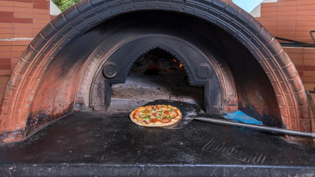 Koka krāsnī cepta pica, kas novietota uz lāpstiņas, gatava izņemšanai. Siltā krāsns iekšpuse ir redzama, ar pelnu un akmeņu tekstūru.