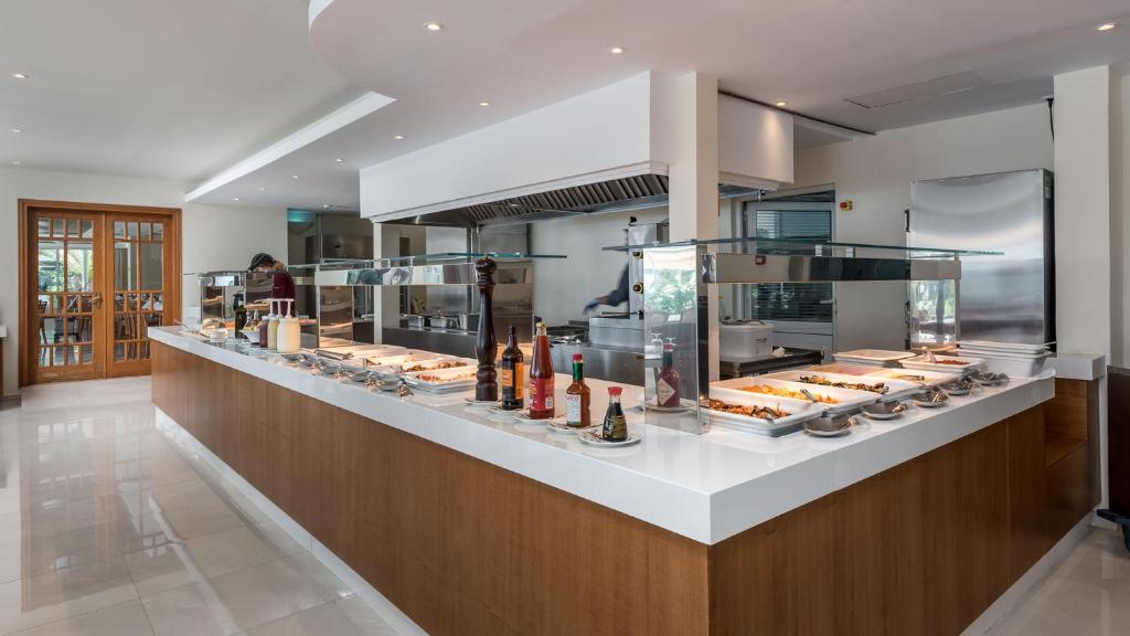 Mūsdienīgs restorāna bufetes galds ar dažādiem ēdieniem un dzērieniem, ko pasniedz uz balta galda. Interjers ir gaišs un elegants, radot patīkamu atmosfēru.