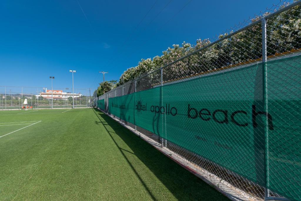 Futbola laukums ar mākslīgo zāli, apjozts ar sietu un reklāmas baneriem, skaidra zila debess fons.