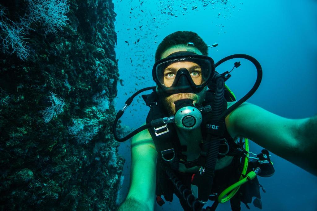 Nirtējs ar elpošanas aparātu, uzņemot pašportretu zemūdens vidē ar ziliem ūdeņiem un koralēm fonā.