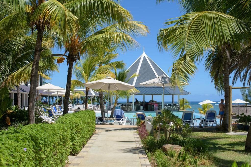 Pastaiga cauri tropu dārziem pie baseina, kur ir saulessargi un atpūtas krēsli, redzams jūras skats un vides relaksējoša atmosfēra.