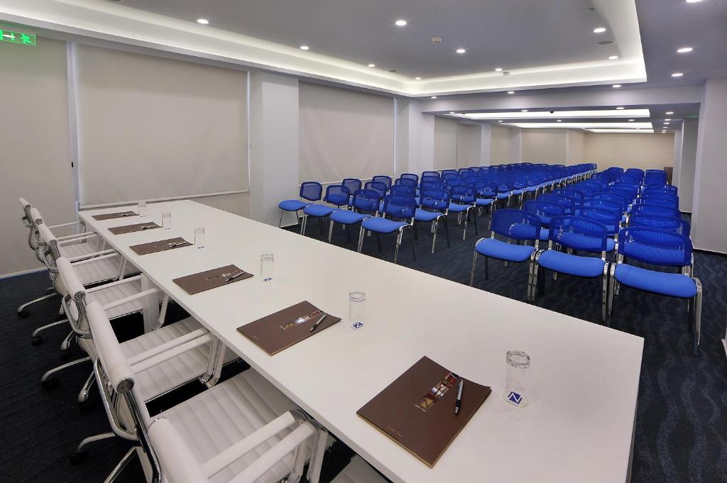 Konferenču telpa ar garu galdu un zilu krēslu rindu. Uz galda ir klātnes un glāzes, radot profesionālu atmosfēru.