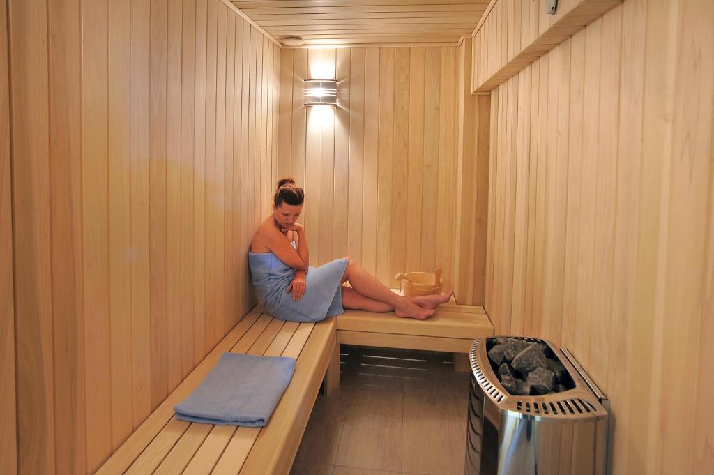 Mūsdienīga sauna ar koka apdari, kurā atpūšas sieviete, apsedzot sevi ar dvieli. Saunā redzama akmens krāsns un koka spainis.