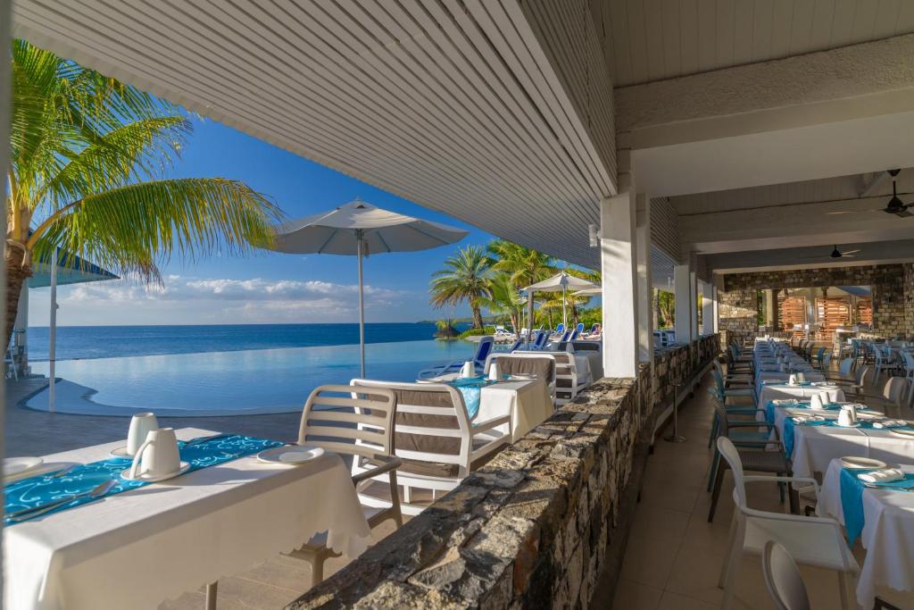 Peldbaseinsā esošais restorāns ar skatu uz jūru un saulainiem palmu kokiem, kas rada relaksējošu atmosfēru. Galdi ir klāti ar baltām galdautiem un dekorēti ar zilu tekstilu.