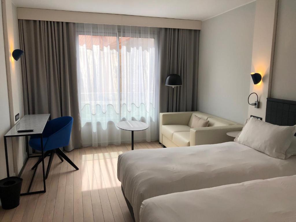 Mūsdienīgs viesnīcas numurs ar divām gultām, gaišām krāsām un lieliem logiem, kas piepildīti ar dabīgu gaismu. Telpā ir dīvāns, maza kafijas galdiņš un rakstāmgalds ar modernu krēslu.