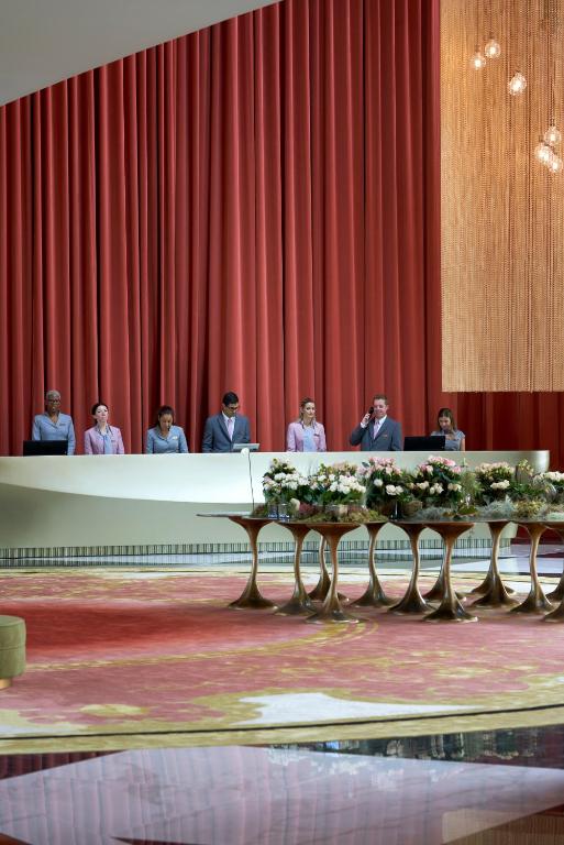 Luksusa viesnīcas reģistratūra ar apkalpojošo personālu un elegantu interjeru. Uz galda izvietoti ziedi, radot viesmīlīgu atmosfēru.