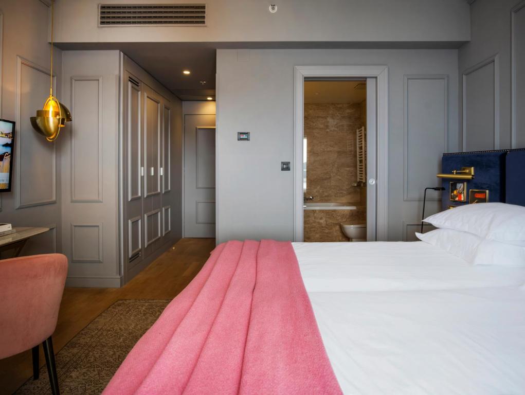 Mūsdienīgs viesnīcas numurs ar elegantu interjeru, kurā redzams gultas veļas komplekts ar rozā segumu, dīvāns un televizors pie sienas. Durvis ved uz vannas istabu, radot mājīgu un komfortablu atmosfēru.