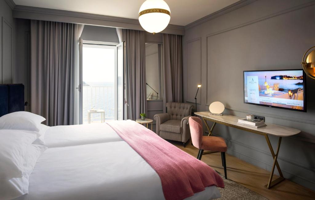 Moderns viesnīcas numurs ar divām gultām, mīkstu rozā segu un skatu uz jūru caur atvērtām durvīm. Numurā ir eleganta mēbeļu izvēle, televizors un mājīgs stūris atpūtas zonai.