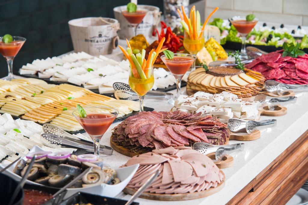 Brokastu bufete ar dažādiem sieriem, gaļas izstrādājumiem un dzērieniem, kas pasniegta elegantā vidē. Krāsaini dārzeņi un garnējumi papildina šo gardo izkārtojumu.
