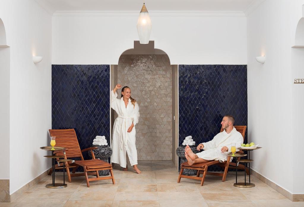 Spa telpa ar diviem cilvēkiem, kuri valkā baltus halātus, relaksējoties uz krēsliem. Uz galda atrodas dzērieni un augļi, radot mierīgu un atpūtu veicinošu atmosfēru.