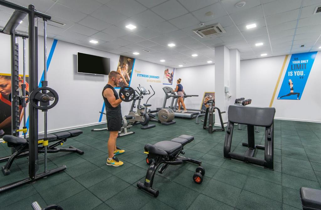 Mūsdienīgs sporta zāles iekārtojums ar trenažieriem un personu, kas veic spēka treniņu. Uz sienas redzami motivējoši uzraksti par fitnesa ceļojumu.