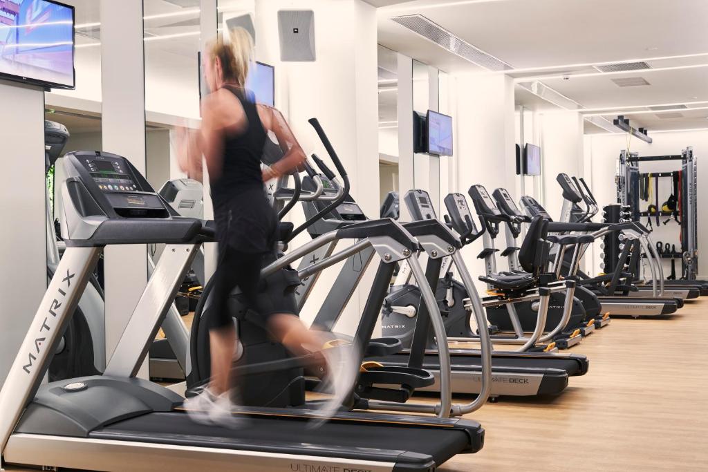 Mūsdienīgs sporta zāles iekārtojums ar skrejceliņu, kurā skrien sportists. Fons ir aprīkots ar vairākiem citiem trenažieriem un televizoriem, kas nodrošina izklaidi treniņa laikā.