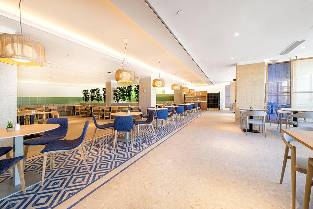 Moderns restorāns ar elegantu interjeru, kas ietver zilu krēslu un ģeometrisku grīdas dizainu. Gaismas ķermeņi piešķir siltumu un atmosfēru telpai.