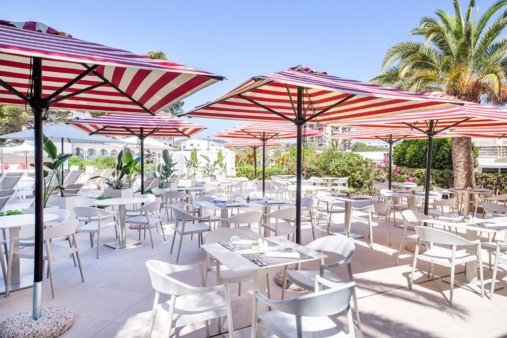 Terases ar sarkano un baltu svītru lietussargiem, kas izvietoti ap galdiem ar baltām mēbelēm, veido patīkamu atmosfēru atpūtai un ēšanai. Apkārt aug zaļumi, radot relaksējošu vidi.