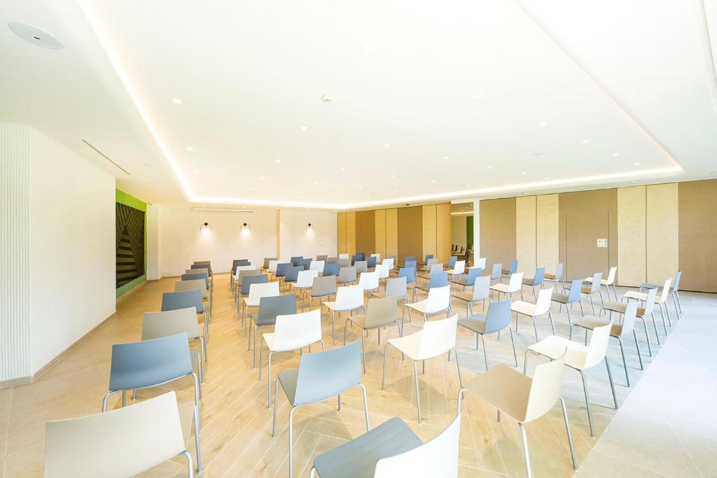 Mūsdienīga konferenču zāle ar rindu sēdvietām, kuras izkārtotas gaišā, plašā telpā ar dabīgu apgaismojumu. Telpa ir aprīkota ar neitrālām krāsām un modernu dizainu.