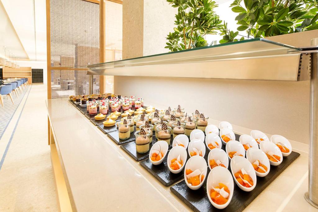 Deserts izvēle ar krāsainiem saldumiem un augļiem, sakārtoti modernā restorānā. Elegants pasniegšanas stils ar zaļumiem fonā.