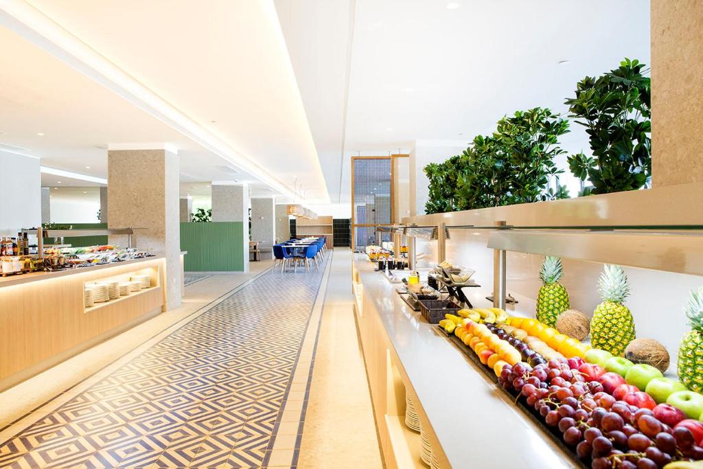 Mūsdienīga restorāna iekšskats ar garu bufeti, kurā redzami dažādi augļi un ēdieni, kā arī stilīgi galdi ar ziliem krēsliem. Gaismas un augi veido patīkamu atmosfēru.