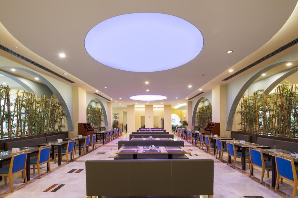 Mūsdienīgs restorāns ar elegantu interjeru, kurā redzami garas galdi un krēsli ar zilu apdari, kā arī liels un apgaismots griestu elements. Zaļumi pie sienām piešķir telpai svaigumu.