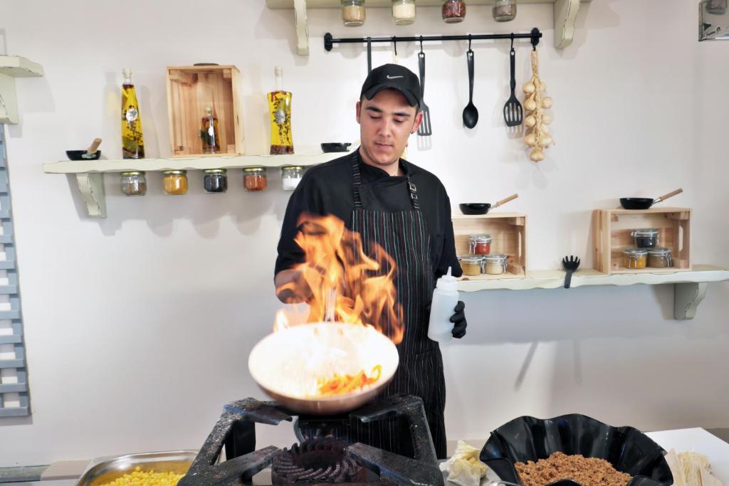 Šefpavārs, kas gatavo ēdienu ar liesmu, aizraujoši strādājot virtuvē, kur redzamas garšvielu burkas un rīku plaukti.